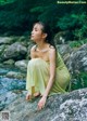 A woman in a yellow dress sitting on a rock by a stream.